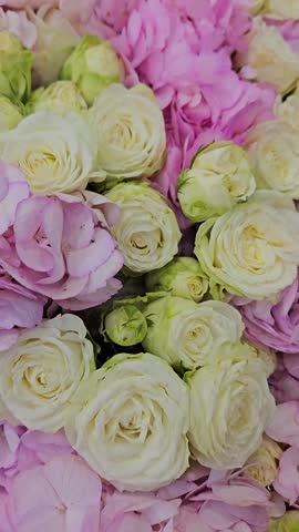 Woman smiles while holding large pink and white floral bouquet. Bouquet contains hydrangeas, roses, and peonies. Flowers are wrapped in soft pink paper. She wears light-colored dress and necklace