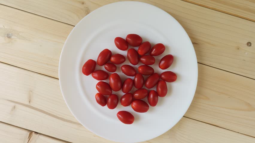Small red ripe grape tomatoes rotating on a white plate on a wooden table, a healthy food concept