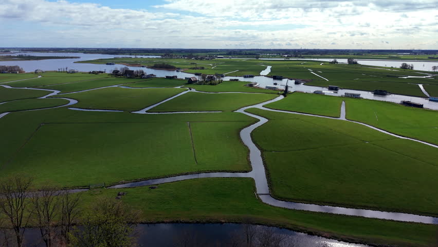 An expansive aerial drone view of a classic Dutch polder landscape, showcasing green fields divided by a network of winding canals and drainage channels under a partly cloudy sky.