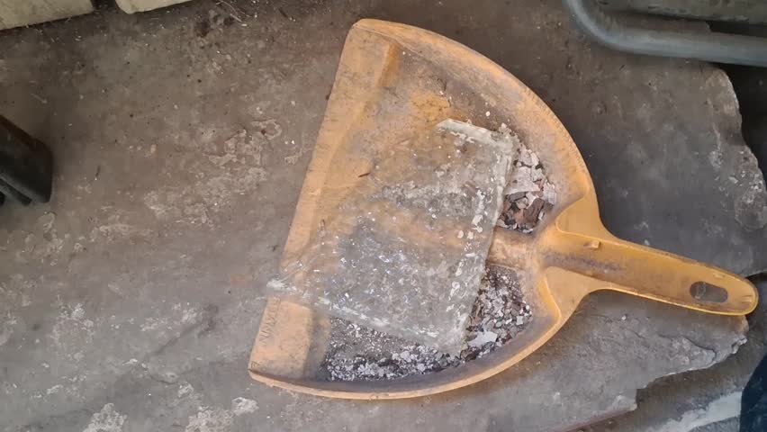 Broken glass sheet rests inside a yellow plastic dustpan while cleaning debris from a construction site in this motion footage.