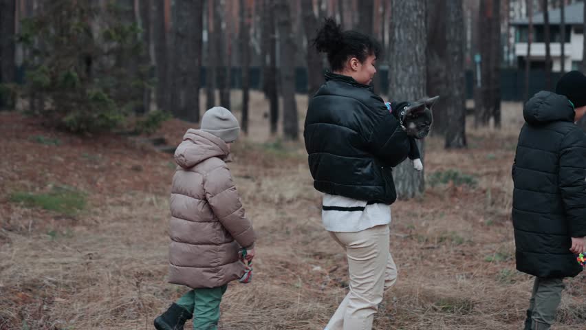 A view from the back of a loving Latino family of three, a mid-20s sister and her two younger brothers, walking in a forest with the sister carrying their French bulldog puppy in her hands