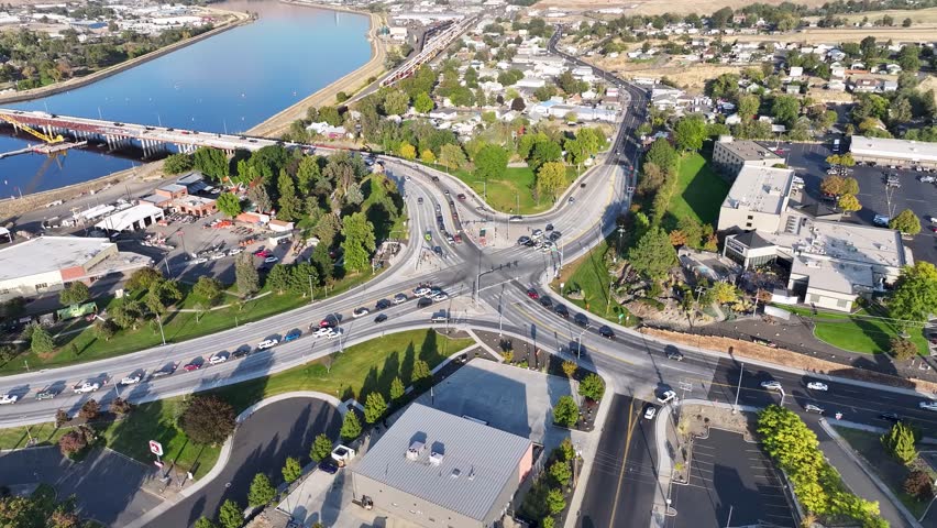 Aerial drone view of a busy urban intersection in Lewiston, Idaho, with the Clearwater River, a lock and dam structure, bridge, and autumn foliage visible across a residential and commercial cityscape on a sunny fall day.