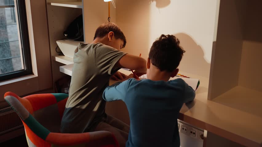 Two multiracial school-aged boys sit at a desk near a window, drawing and painting under a desk lamp in a modern home study area.