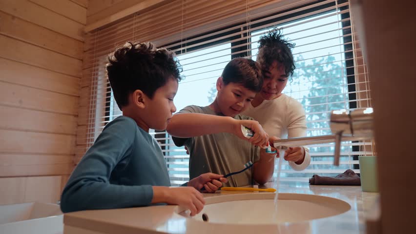 The son stands and applies toothpaste to the toothbrushes of his mother and his brother who are standing next to him, then applies it to himself