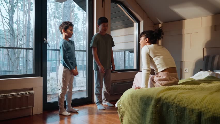 A teenage Latino boy with short black hair, talking enthusiastically to his older, mid-20s sister, as he stands next to the bed where she sits in a cozy bedroom, with his younger brother beside him
