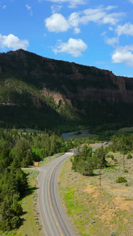 Vertical Screen: Scenic Montana highway winds through dramatic mountain valley with towering rocky cliffs, winding river, lush green pine forests, and white truck traveling under bright blue sky.
