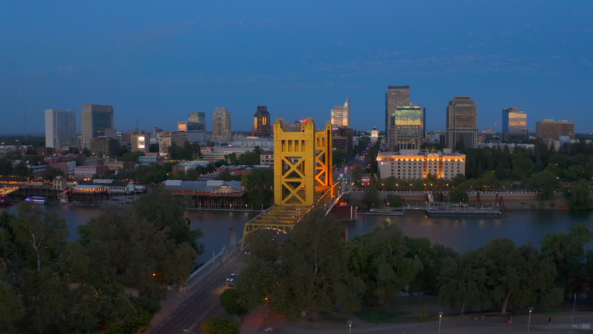 Aerial drone footage of Sacramento skyline at blue hour featuring the illuminated Tower Bridge crossing the Sacramento River with downtown city lights glowing at dusk.