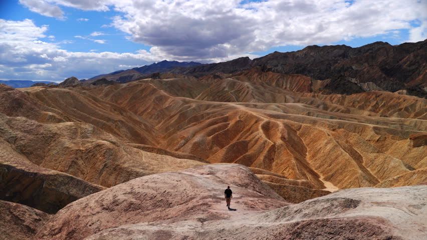 Man explores colorful eroded badlands at Zabriskie Point in Death Valley National Park. Golden and orange sedimentary rock formations stretch toward distant mountains under dramatic cloudy sky.