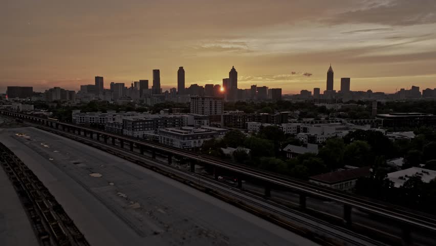 Dynamic aerial footage captures Atlanta city skyline at sunset, showcasing towering buildings silhouetted against vivid sky, with metro train passing below. Cinematic clip highlights vibrant urban landscape at dusk.