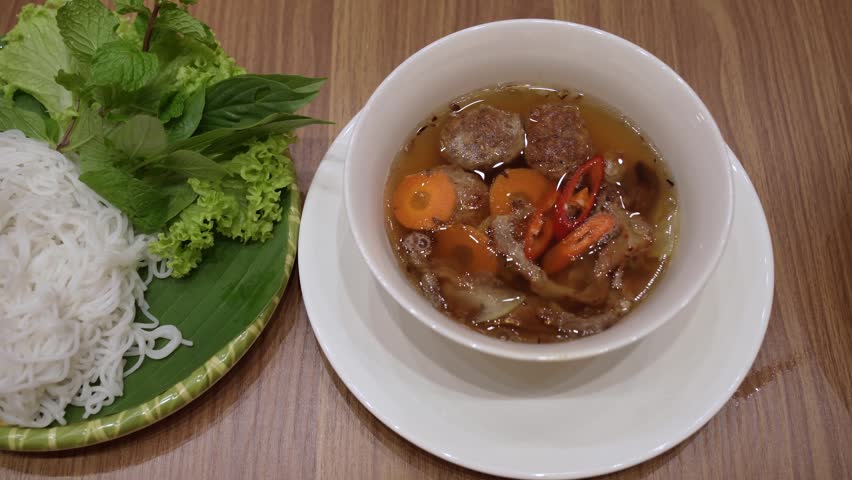 Bun Cha is a classic Vietnamese dish featuring grilled pork served in a savory-sweet dipping broth with pickled vegetables. In this image, a bowl of grilled pork slices and meatballs in broth is accompanied by a plate of fresh rice vermicelli noodles, lettuce, and aromatic herbs.