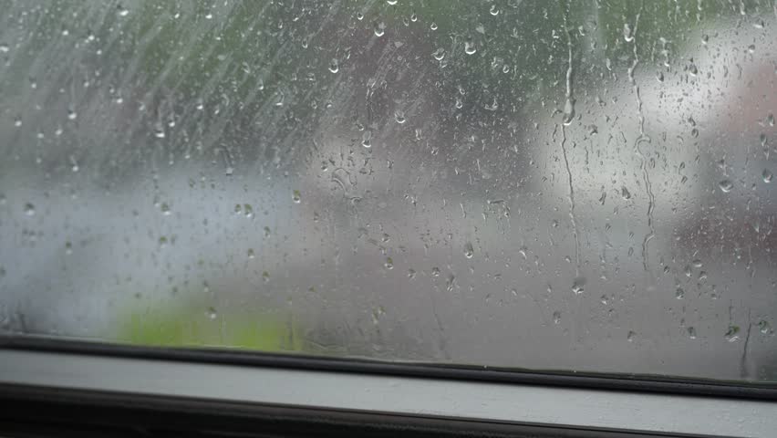 Raindrops resting on a window pane during a rainy day, with a softly blurred outdoor background visible through the glass. The scene captures a calm, peaceful atmosphere and the cozy feeling of staying indoors while watching the rain outside