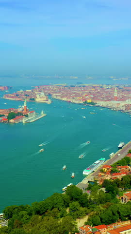 Vertical video. Venice, Italy. A sweeping aerial panoramic view captures the historic center of Venice spread across its lagoon archipelago under a clear blue sky. The footage reveals the dense urban fabric of the city, with countless terracotta rooftops forming a warm, textured carpet that contrasts sharply with the deep turquoise waters of the Venetian Lagoon. The camera glides over the Grand Canal and surrounding waterways, where boats and watercraft trace white wakes across the surface. Key landmarks punctuate the skyline, including the distinctive silhouette of St Mark