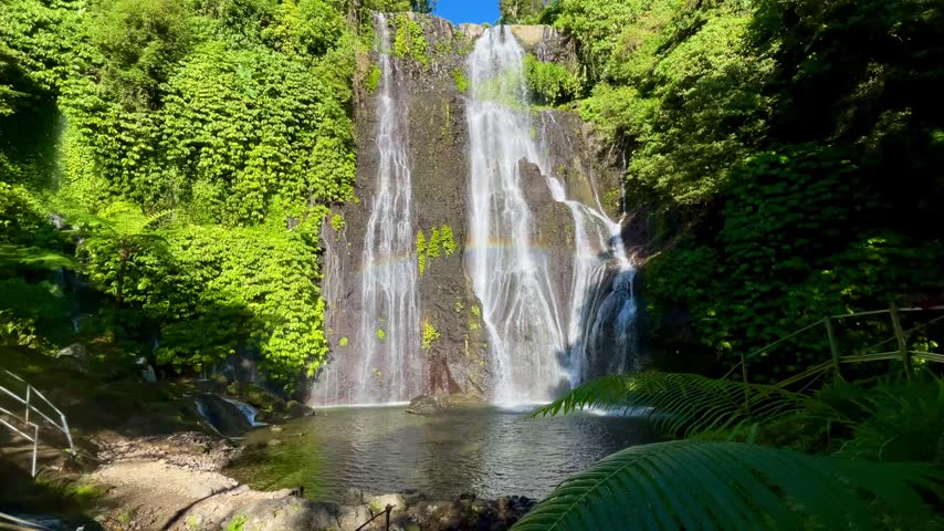 A beautiful wide angle horizontal static establishing shot of a majestic twin waterfall with a vibrant natural rainbow over a clear natural pool under a bright sunny sky in Bali