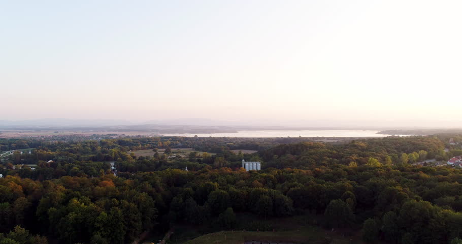 Elevated view of a wooded landscape at sunrise with treetops filling the foreground and a calm lake visible on the horizon under a bright morning sky