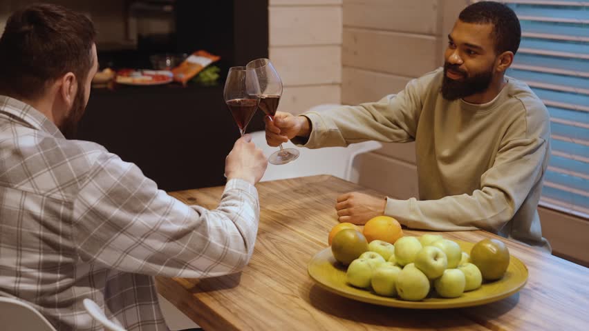 A Black man in a beige sweatshirt and a White man in a plaid shirt in their 30s, sit facing each other at a wooden dining table with a big plate of fruits at home, clink glasses of red wine and talk.