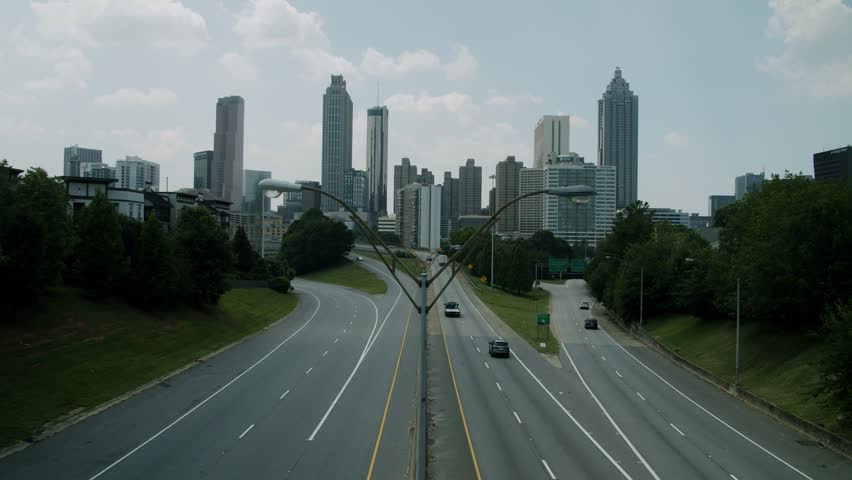 Wide shot showcasing urban landscape with skyscrapers and moving vehicles on asphalt roads under white clouds. Iconic city buildings stand tall behind busy expressway in Georgia state capital.