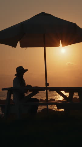 A woman works on her laptop at a beachside table under an umbrella during a beautiful sunset. This is a vertical video.