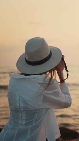 A photographer wearing a hat takes a photo of the sunset over the ocean. Vertical video.
