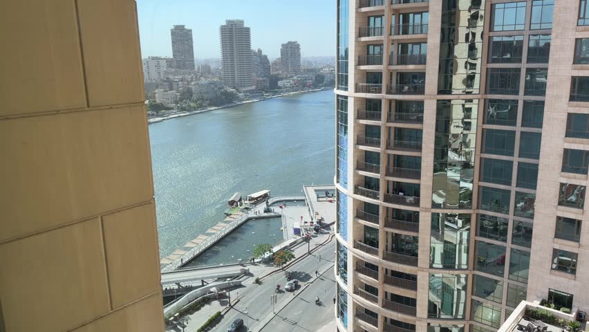 A scenic high-angle video of the majestic Nile River in Cairo, Egypt, featuring modern high-rise buildings, boats on the water, and a waterfront street with moving traffic under a clear blue sky on a sunny day.