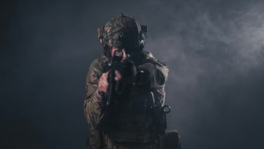 Soldier in military uniform points firearm at the viewer, demonstrating readiness and concentration in a low-light, smoky setting