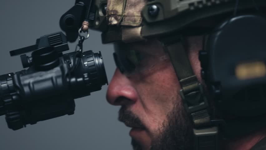 Male soldier in tactical gear carefully aligns night vision scope on rifle, demonstrating precision and focus in a low-light indoor setting