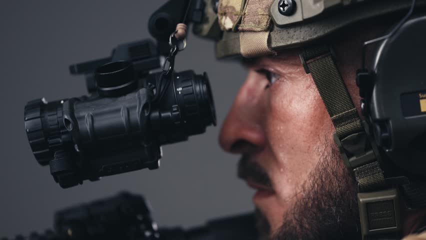 Soldier wearing tactical helmet aims through night vision scope, displaying concentration and readiness in a neutral indoor setting with soft lighting