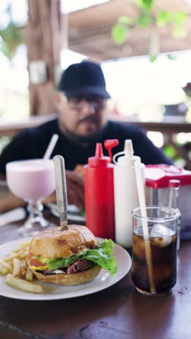 Close up of a complete meal with a cheeseburger, french fries, soda, and milkshake. A man enjoying his food in the blurred background
