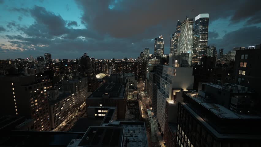 Dramatic aerial shot capturing urban architecture in New York City during twilight hour. Vibrant city lights illuminate tall buildings while dark storm clouds drift across deep blue sky above.
