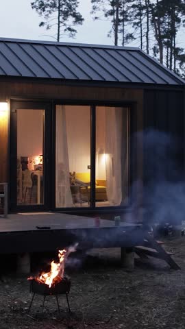 Vertical video. Modern cabin with illuminated windows and outdoor fire pit at dusk in forest.