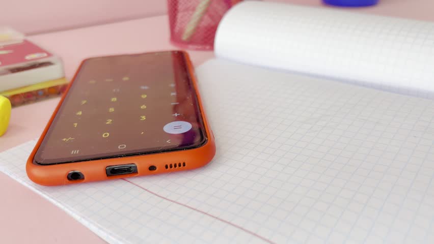 Student using smartphone calculator for math homework with a pencil and notebook on a pink desk, symbolizing modern education, distance learning, and academic study