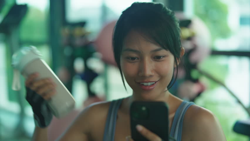 Asian woman in gray sportswear smiles at her phone while holding a protein shaker bottle during a break in a bright gym.