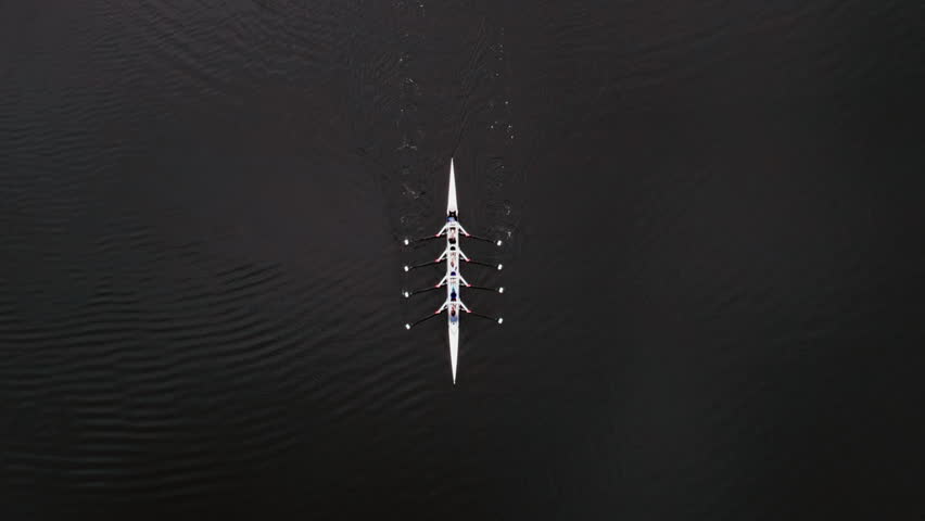 Aerial drone view of a rowing team moving oars in perfect sync during training. Overhead shot captures rhythmic motion, symmetrical foam trails, and glassy river reflections.