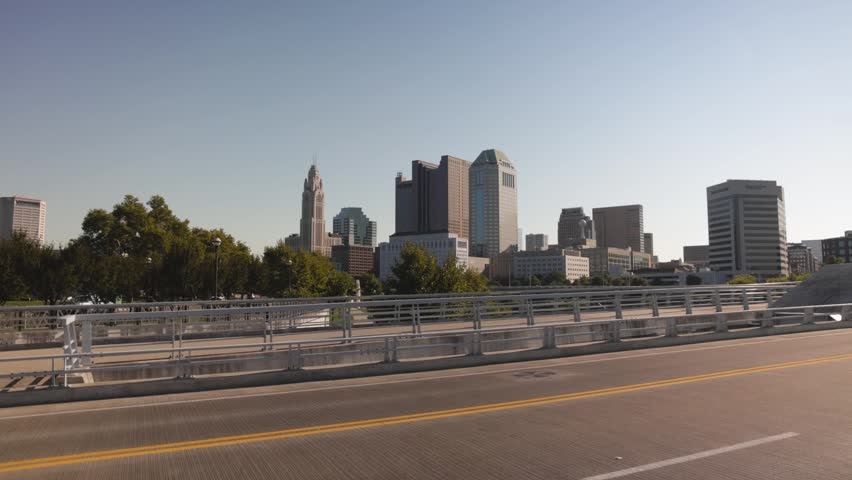 Dynamic motion shot crossing urban bridge reveals downtown Columbus Ohio city skyline. Cinematic footage of metropolitan landscape featuring LeVeque Tower and corporate office architecture.
