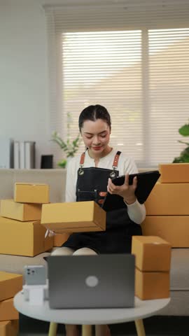 Young Asian woman running a small e commerce business, checking inventory and preparing cardboard boxes for shipping from her living room