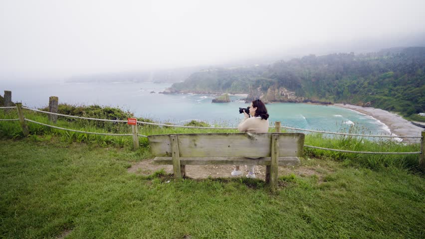 Young female content creator sitting on a wooden bench. Capturing the dramatic coastal scenery with a professional camera during a foggy day. Enjoying a moment of quiet contemplation and inspiration