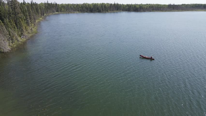 Drone flying over a person in a red canoe while floating on a small northern lake that is surrounded by an evergreen forest