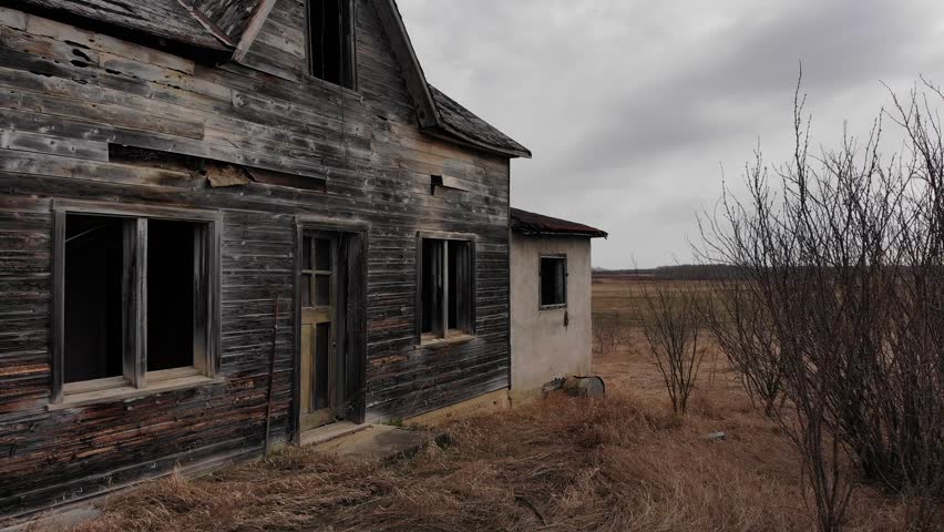 Moving sideways looking at an abandoned wooden prairie farmhouse with the windows missing on a cloudy day