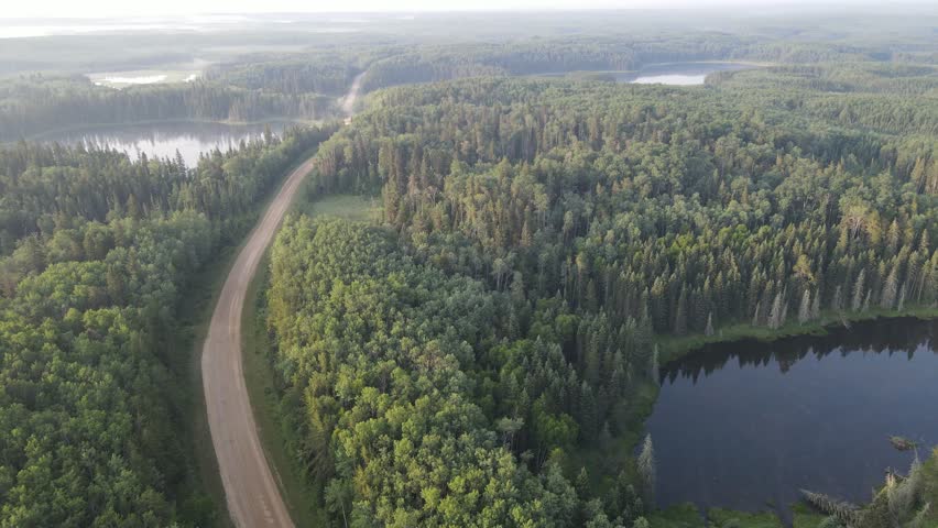 Drone view looking down at a gravel road winding through a northern park forest with many small lakes. The distant trees are are hidden in forest fire smoke.