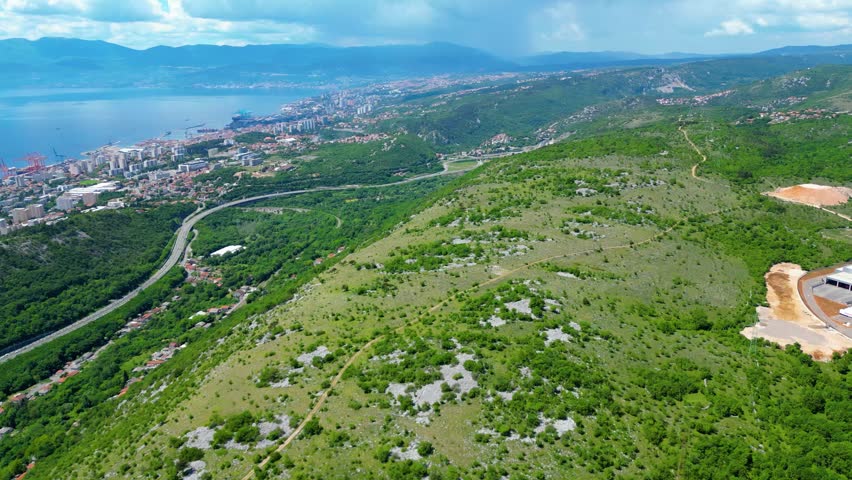 An expansive aerial view of Rijeka, Croatia, showcasing the vibrant coastal city, bustling port, and winding highways amidst lush green hills and the serene Kvarner Bay.