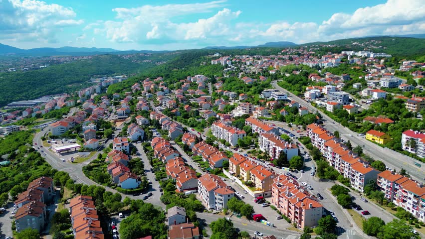 Aerial view of a vibrant residential area in Rijeka, Croatia, nestled on green hills under a bright blue sky with scattered clouds. Terracotta roofs dot the landscape. Skurinje