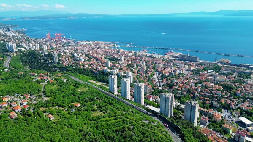 Aerial view of Rijeka, Croatia, showcasing its vibrant port, urban sprawl, and lush green hills meeting the clear blue Adriatic Sea under a bright sky. A dynamic coastal landscape. Skyscrappers on Banderovo