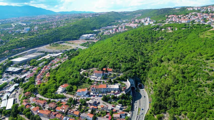 Aerial view of Rijeka, Croatia, showcasing its vibrant urban landscape, lush green hills, and the sparkling Adriatic Sea under a clear sky. A modern highway connects the city.