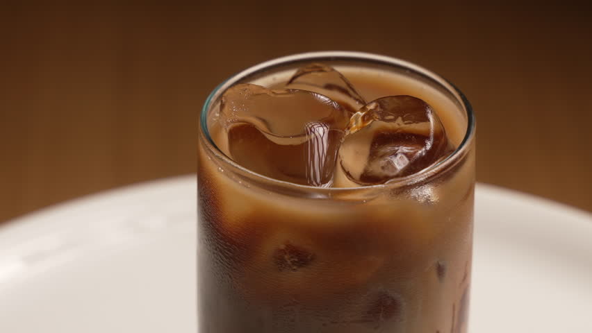 Close up of a cold brew coffee with milk and ice cubes spinning in a glass on a white plate, fa01