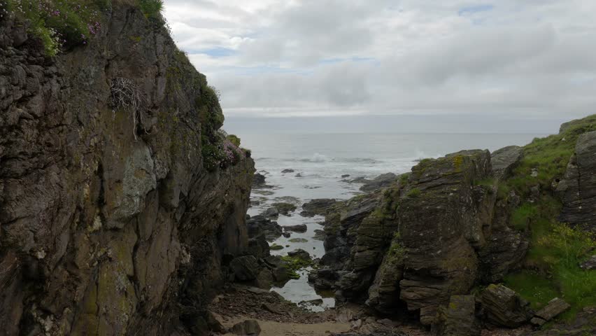 The drone slowly glides over the rugged coastline of Kintyre in Scotland. At first, the frame shows a narrow rocky cleft between two tall cliffs covered in moss, grass, and scattered pinkish-purple flowers. The camera seems to hover over this natural corridor, and beyond it opens a view of the calm grey sea.
Then the drone shifts slightly forward and higher, and the cliffs spread apart: on the left, a steep wall with a rough, wet stone texture; on the right, a layered outcrop overgrown with greenery. Below, between them, you can see dark boulders and small pools of seawater left behind by the tide. Everything feels cold, fresh, and very quiet.
Next, the camera emerges toward the open shore, and the landscape fully раскрывается: in front of it stretches a wide ocean dotted with black reefs rising out of the water. Waves gently roll across the rocks, leaving a thin line of white foam. The horizon is almost level, and the sky is low and heavy, covered with pale grey clouds, though faint breaks in the cloud cover let a little light through.
Then the drone pulls back slightly and shows the shoreline relief from another angle: on both sides are powerful rock formations covered in vivid green moss and grass, while in between lie shallow tidal pools and scattered stones. The sea surface looks metallic grey, calm, with only a faint ripple. The movement in the frame is very slow, almost meditative.
In the final moments, the camera seems to move slightly closer to one of the cliffs, and a steep rock face fills the foreground, densely covered with greenery and layered stone. Behind it are the endless sea, the cold sky, and the hazy line of the horizon. There are no people in the shot — only nature, stone, and wind — creating a feeling of solitude, silence, and the ancient, almost primal beauty of the Scottish coast.