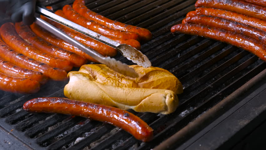 Delicious sausages and hot dog buns cooking on the grill. Juicy sausages and bread buns grilling over hot charcoal at a food festival. A street food vendor uses metal tongs to turn the food
