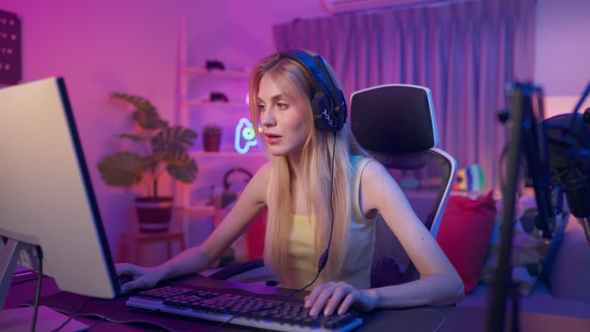 Caucasian gamer wearing headphones, playing online video game on desktop. Attractive young female player sit at desk, enjoying live gaming, esports, and digital entertainment in colorful neon room.