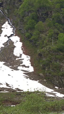 A vertical tilt shot reveals a waterfall cascading down rocky cliffs amidst melting spring snow.