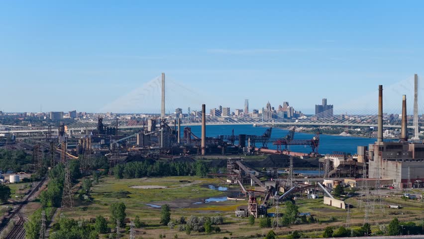 Aerial view of a large industrial complex with the Detroit skyline and Gordie Howe Bridge. in the background.