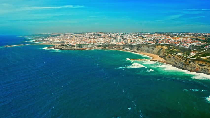 Drone is panning   European touristic town from the ocean.   Drone aerial view over beach, coastline in Ericeira, Portugal, on summer sunny day. Beautiful cityscape with skyline, ocean rocky shore  