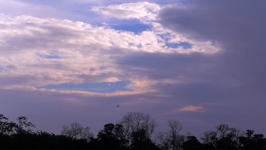 Layered clouds drift across the sky above a rural landscape, revealing patches of blue between the formations. The scene captures the beauty of changing weather and natural light in the countryside.
